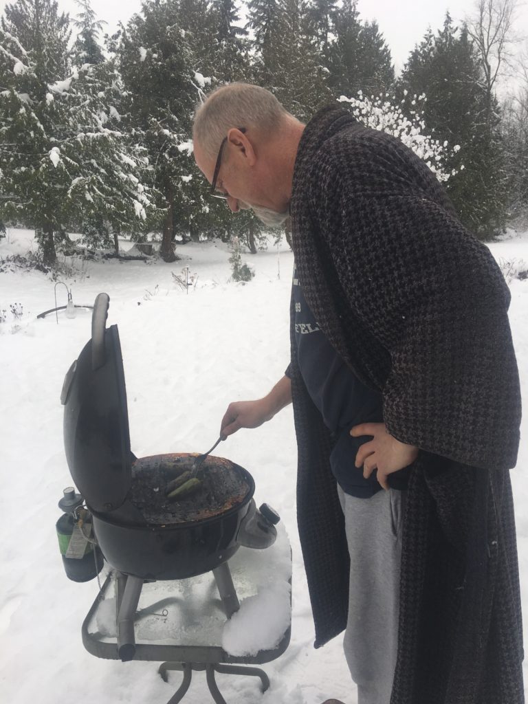 Man in a robe bent over a grill in the middle of a snowstorm.