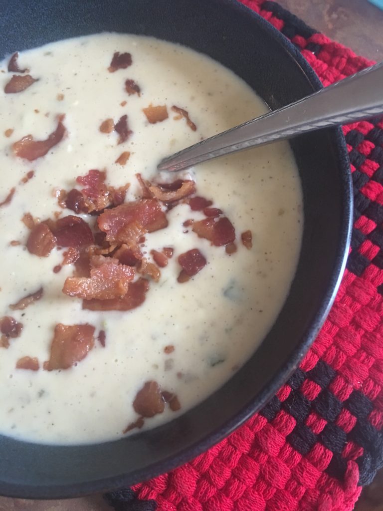 Bowl of soup on red and black checkered cloth, with bacon bits scattered over top and spoon resting in center